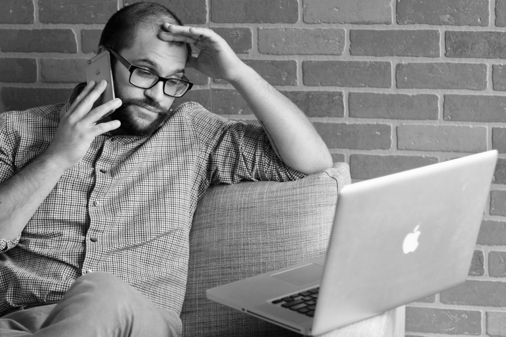 A man in glasses on a phone call, looking worried, sitting next to a laptop in an office