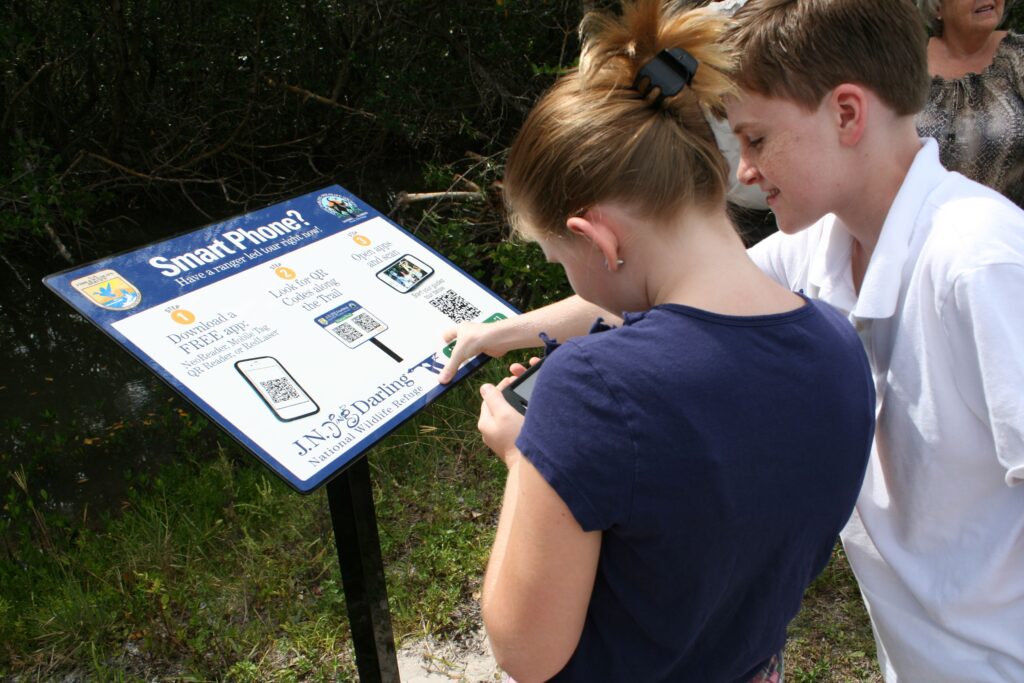 Two visitors hold up their smartphones to scan QR codes on signs along an outdoor nature trail